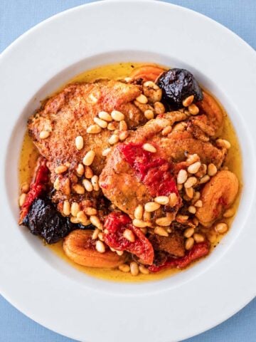 Braised Chicken Thighs with Prunes and Apricots served in a white bowl, topped with toasted pine nuts and simmered in a rich sauce with tender dried fruit.