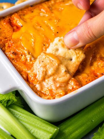 Hand dipping a cracker to scoop Rotisserie Buffalo Chicken Dip with melted cheddar