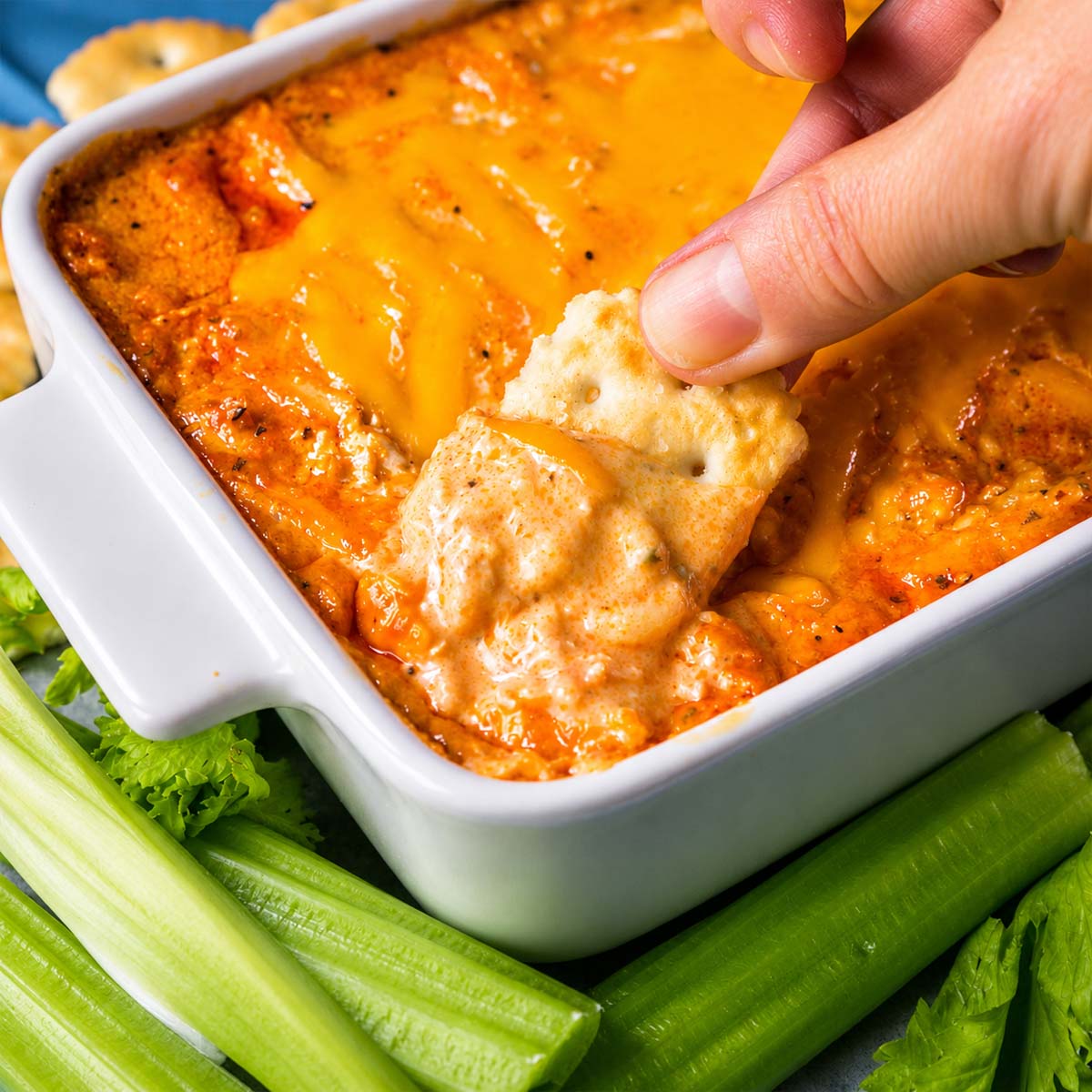 Hand dipping a cracker to scoop Rotisserie Buffalo Chicken Dip with melted cheddar