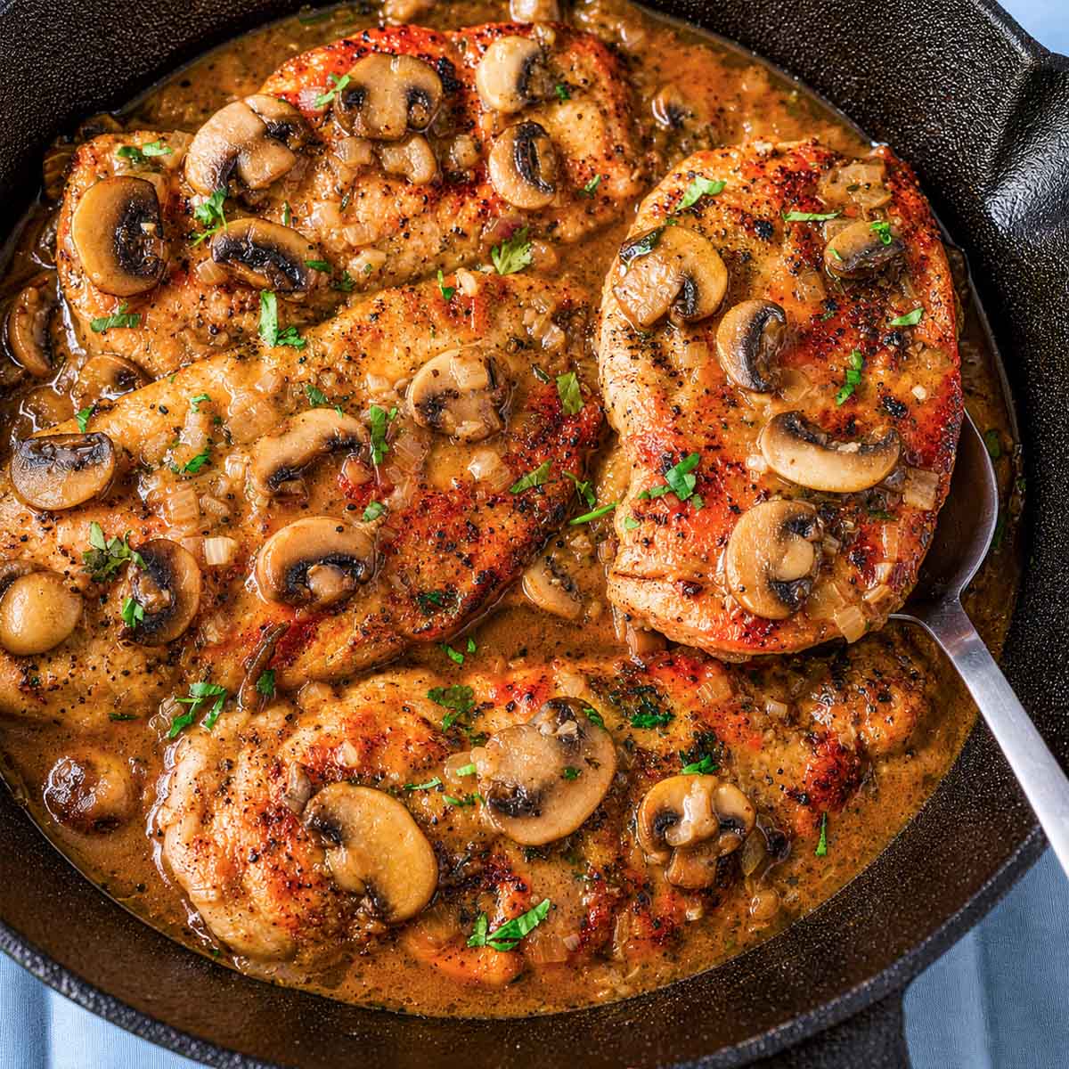Chicken Marsala without cream in a cast iron skillet with sliced mushrooms, onions, and Marsala wine sauce, with one chicken cutlet lifted on a spoon.
