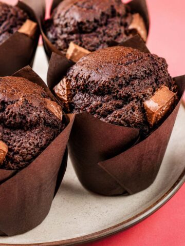 Chocolate muffins in brown tulip liners on a white plate, with chocolate pieces baked into the sides, against a pink background.