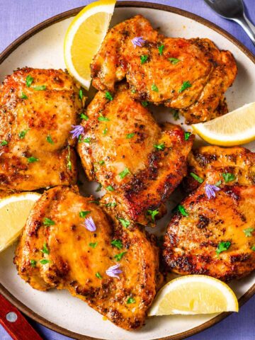 Greek chicken thighs with golden, herb-marinated meat and lemon wedges on a white plate, ready to serve as an easy Mediterranean dinner.