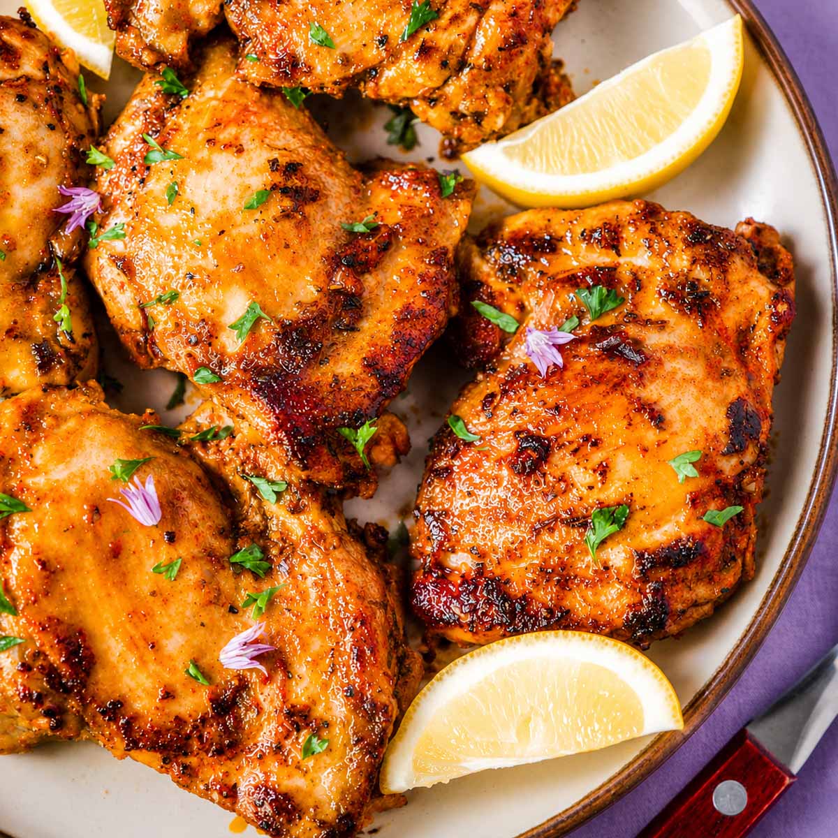 Close-up of juicy Greek chicken thighs with a golden garlic and herb marinade, garnished with parsley and lemon wedges on a plate.