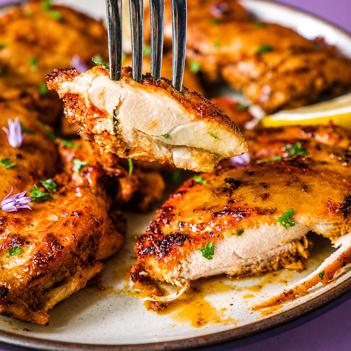 Fork holding a juicy piece of Greek chicken thigh, showing the tender inside and golden garlic-herb crust, with more marinated chicken on a plate in the background.