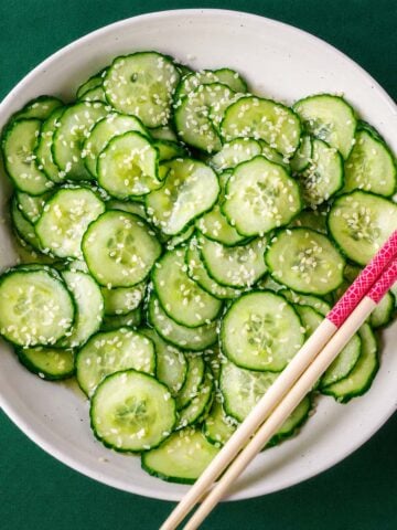 Japanese cucumber salad (sunomono) with thinly sliced cucumbers and sesame seeds in a white bowl, served with chopsticks on a green background.