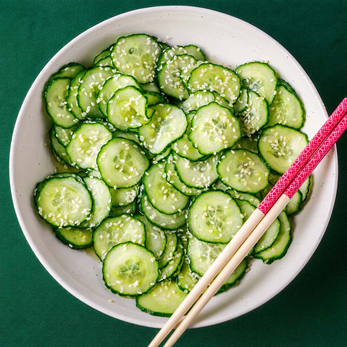 Japanese cucumber salad (sunomono) with thinly sliced cucumbers and sesame seeds in a white bowl, served with chopsticks on a green background.