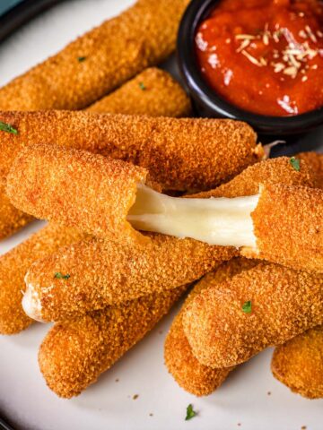 Crispy homemade mozzarella sticks stacked on a plate, with one pulled apart to show the gooey melted cheese and a small bowl of marinara sauce for dipping.