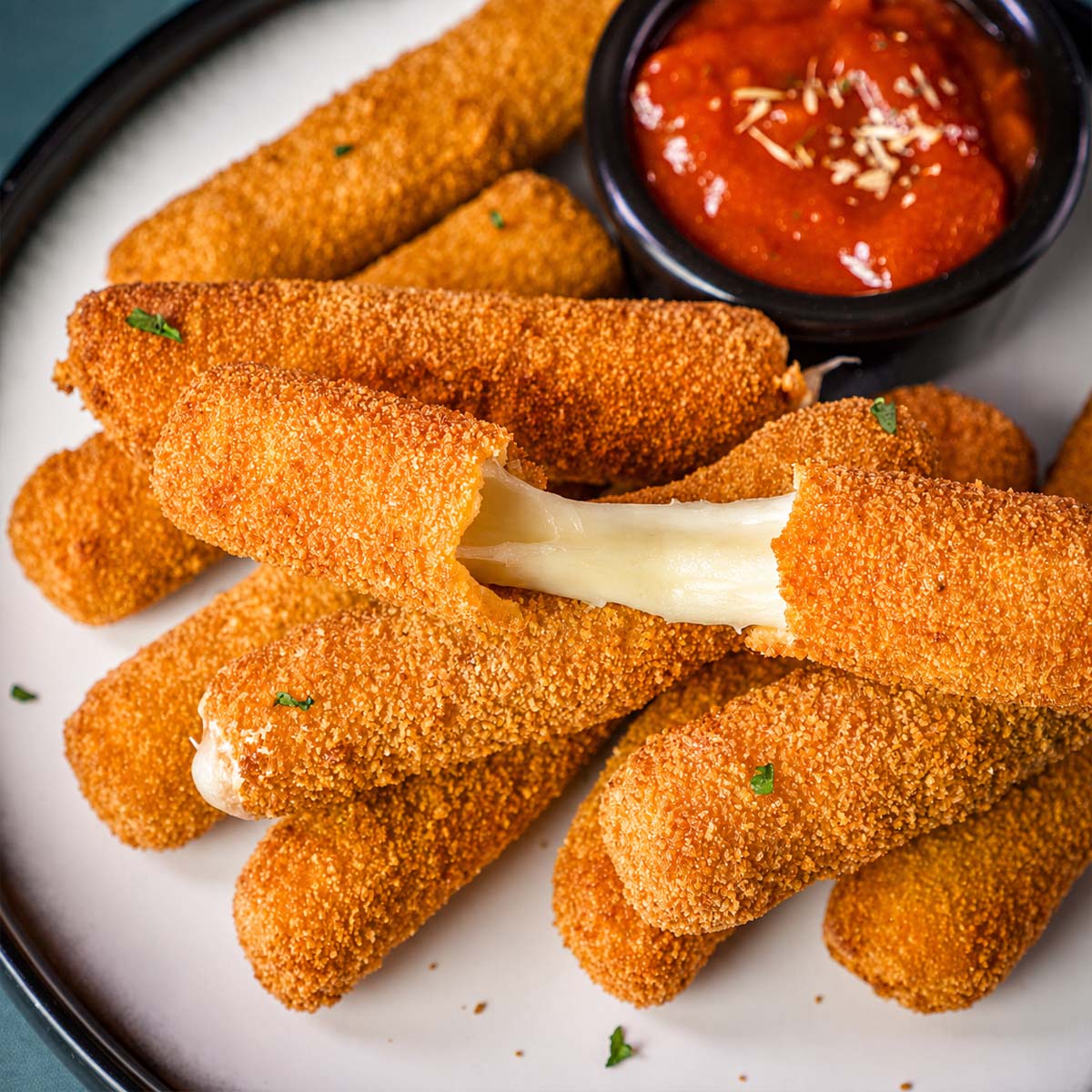 Crispy homemade mozzarella sticks stacked on a plate, with one pulled apart to show the gooey melted cheese and a small bowl of marinara sauce for dipping.
