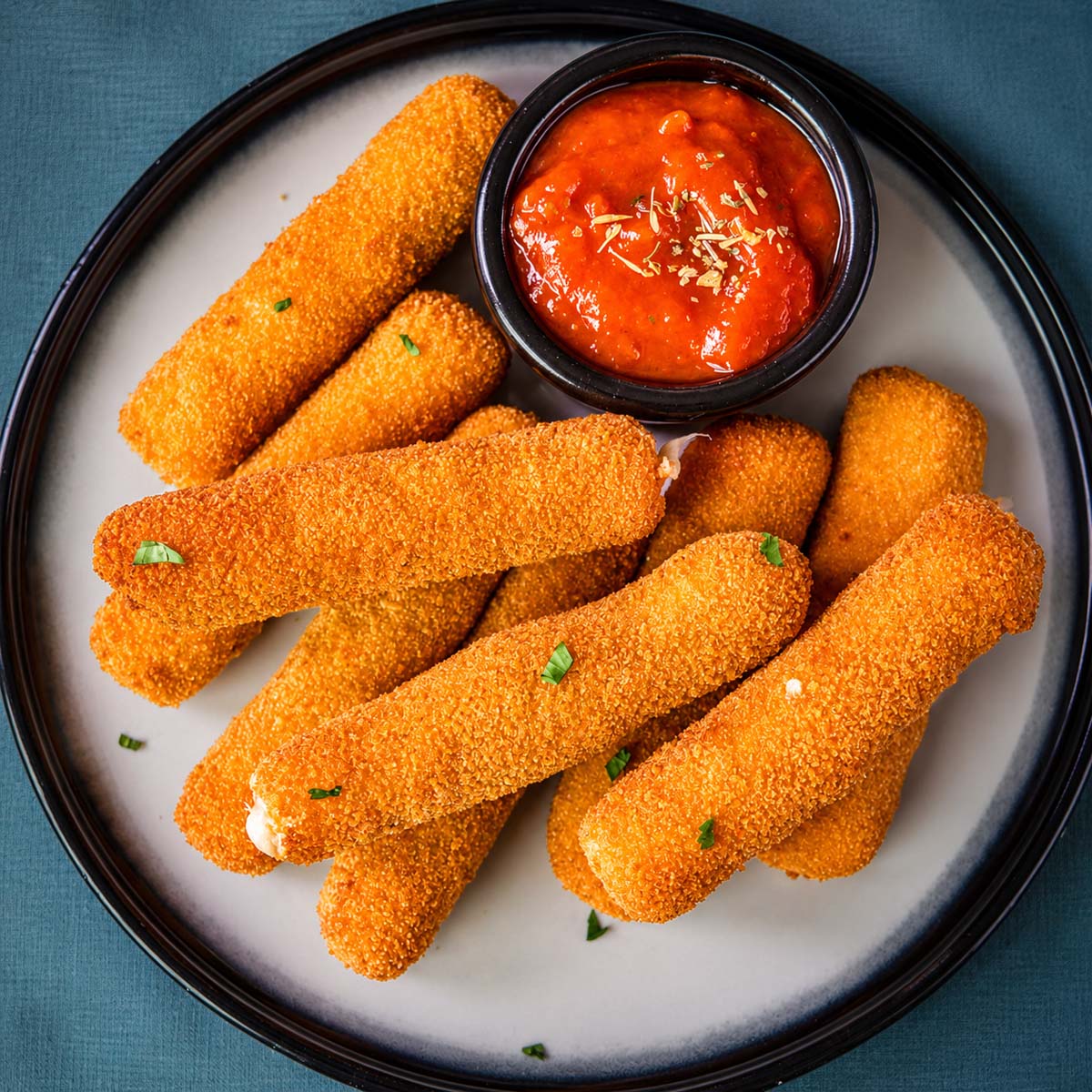 A plate of golden, crispy homemade mozzarella sticks served with a small bowl of marinara sauce for dipping.