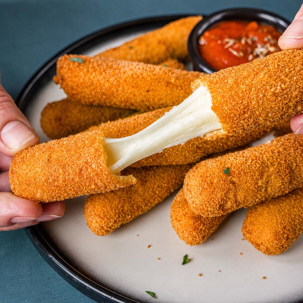 A close-up of two hands pulling apart a crispy homemade mozzarella stick, revealing the gooey melted cheese inside, with more mozzarella sticks on the plate.