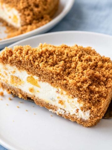 One serving of a Peach cheesecake slice with crumb topping on a white plate, featuring a creamy no-bake filling and peach pieces