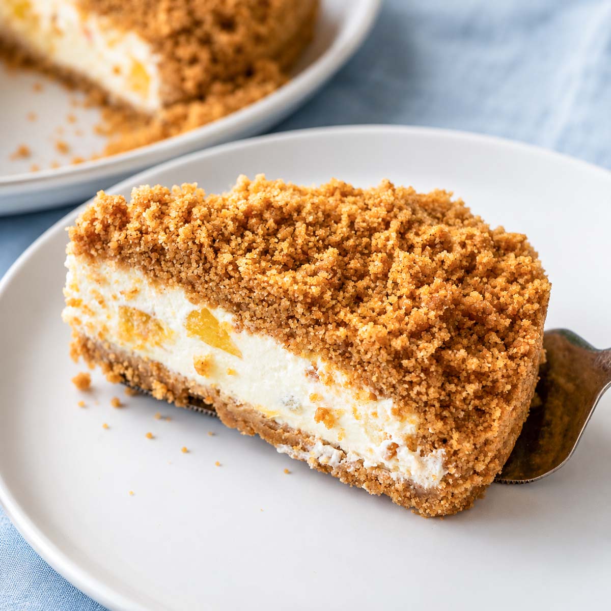 One serving of a Peach cheesecake slice with crumb topping on a white plate, featuring a creamy no-bake filling and peach pieces