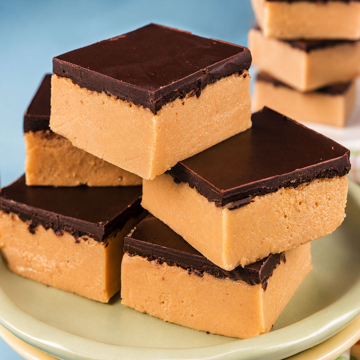 Close up of peanut butter bars with a smooth dark chocolate topping stacked on a plate