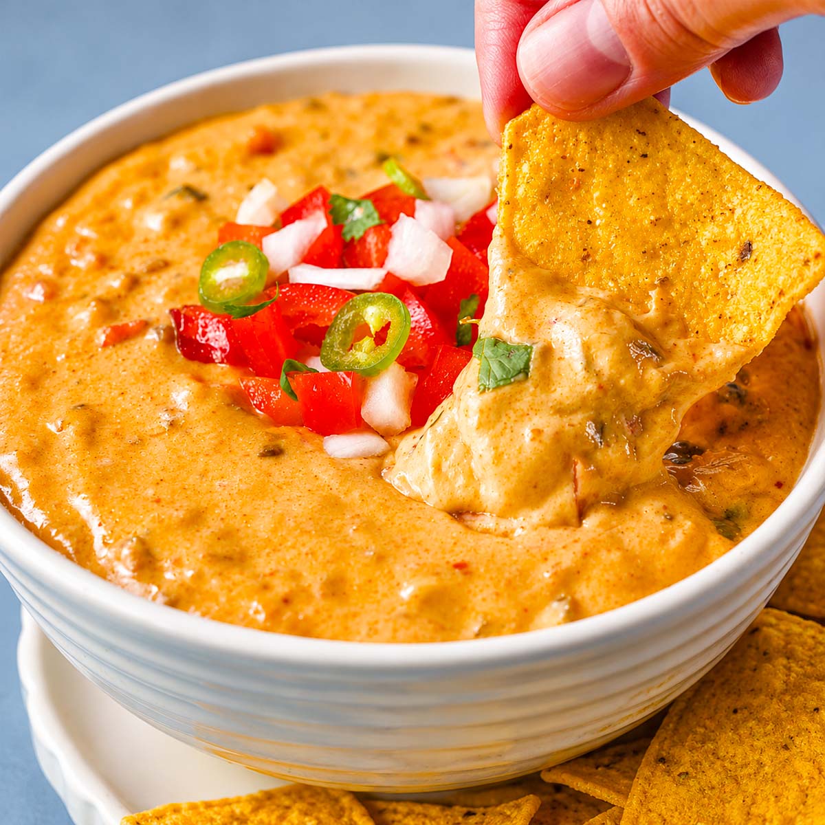 Tortilla chip dipping into creamy queso dip topped with pico de gallo and jalapeño slices.