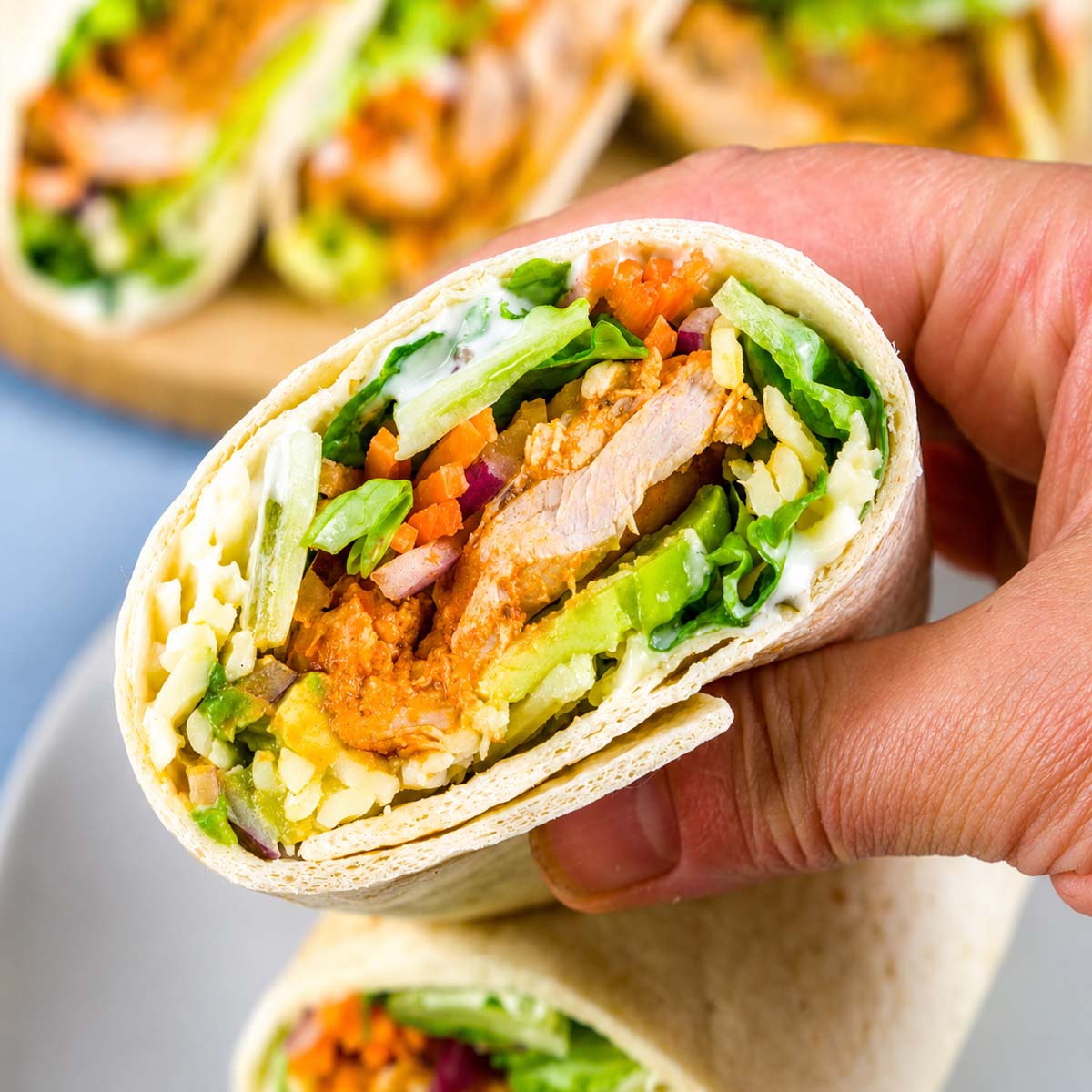 Close-up of a hand holding a buffalo chicken wrap filled with spicy chicken, avocado, lettuce, carrots, and cheese