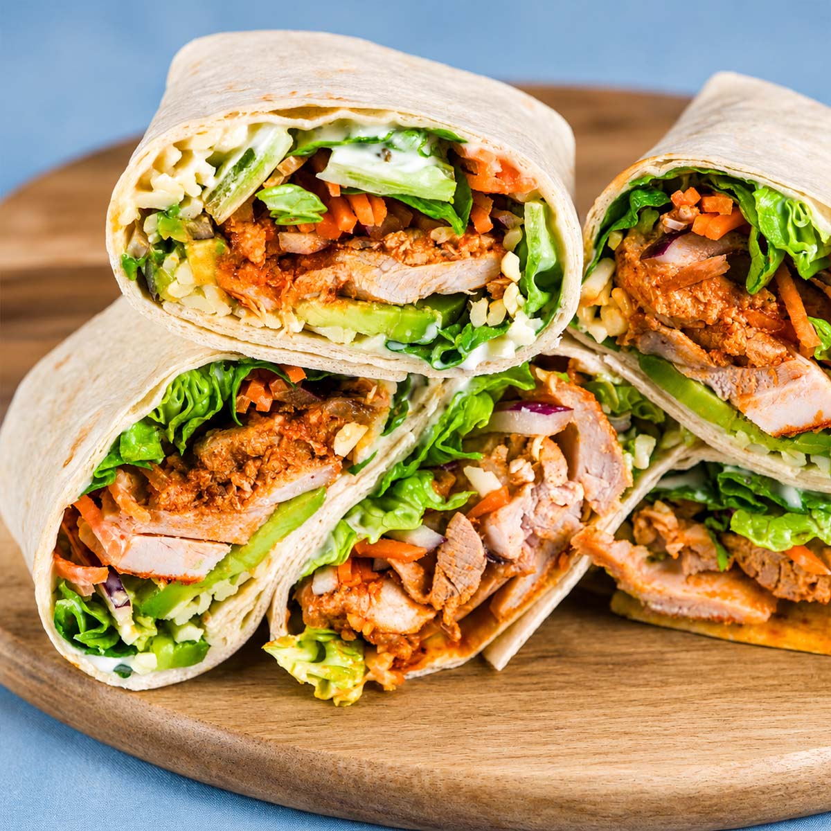 Stacked buffalo chicken wraps on a wooden board with tender chicken, crisp lettuce, avocado, carrots, and ranch dressing