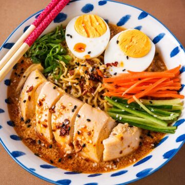 Bowl of chicken ramen noodle soup with sliced chicken breast, ramen noodles, soft-boiled eggs, shredded carrots, cucumber strips, green onions, and sesame seeds in a creamy spicy peanut broth.