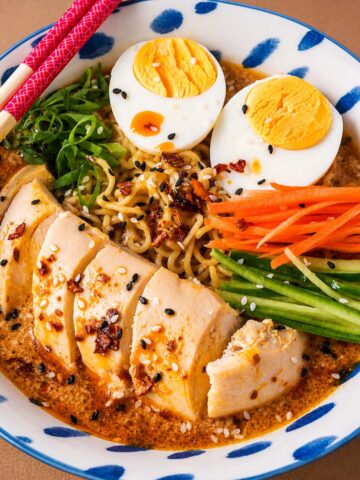 Bowl of chicken ramen noodle soup with sliced chicken breast, ramen noodles, soft-boiled eggs, shredded carrots, cucumber strips, green onions, and sesame seeds in a creamy spicy peanut broth.