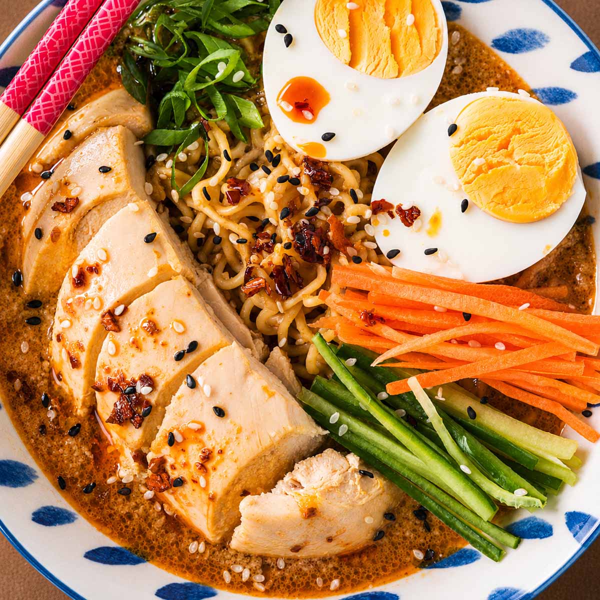 Close-up of chicken ramen noodle soup with sliced chicken breast, ramen noodles, boiled eggs, shredded carrots, cucumber strips, green onions, and sesame seeds in a creamy spicy broth.