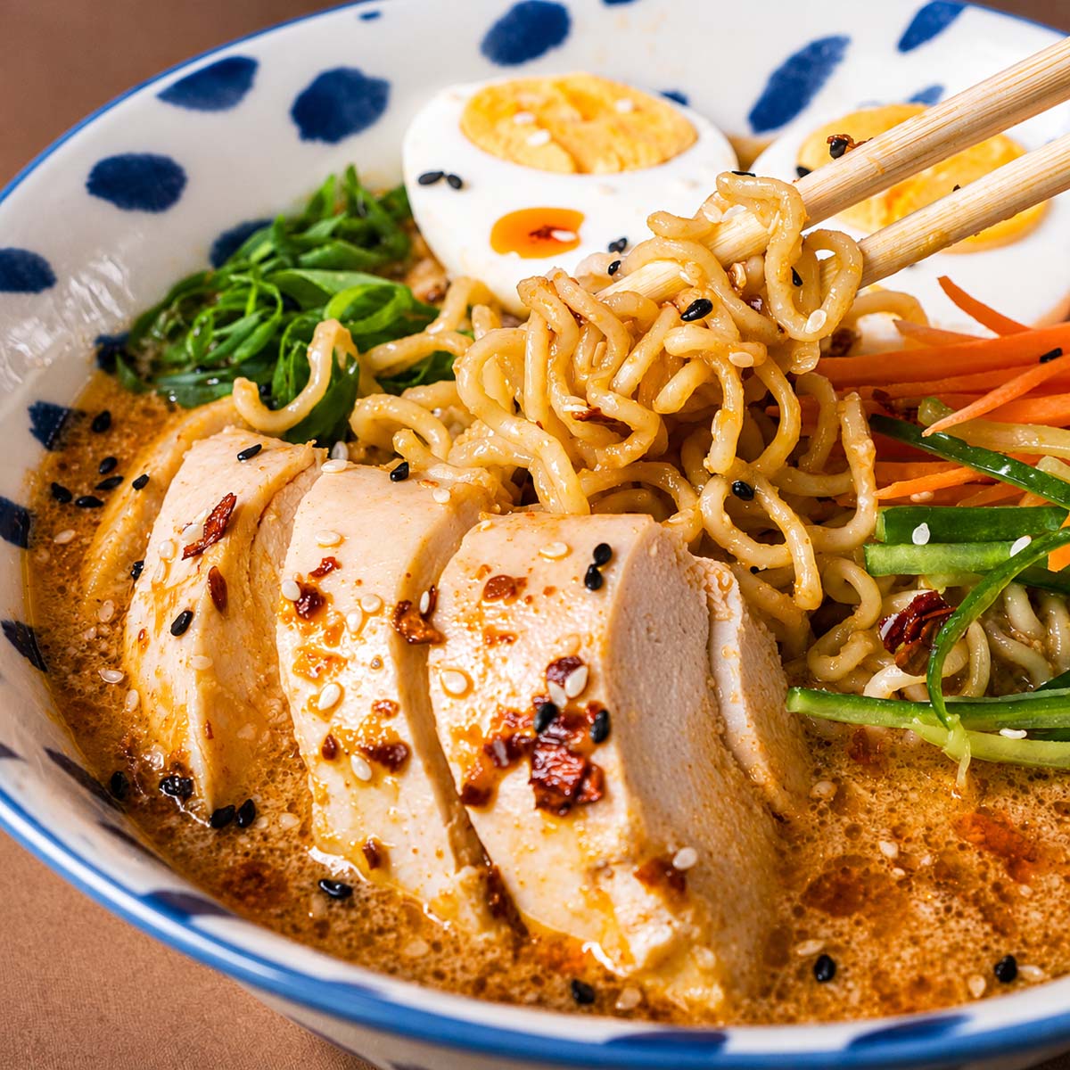 Close-up of chicken ramen noodle soup as chopsticks lift the noodles, showing sliced chicken breast, boiled eggs, shredded vegetables, and sesame seeds in a creamy spicy broth.