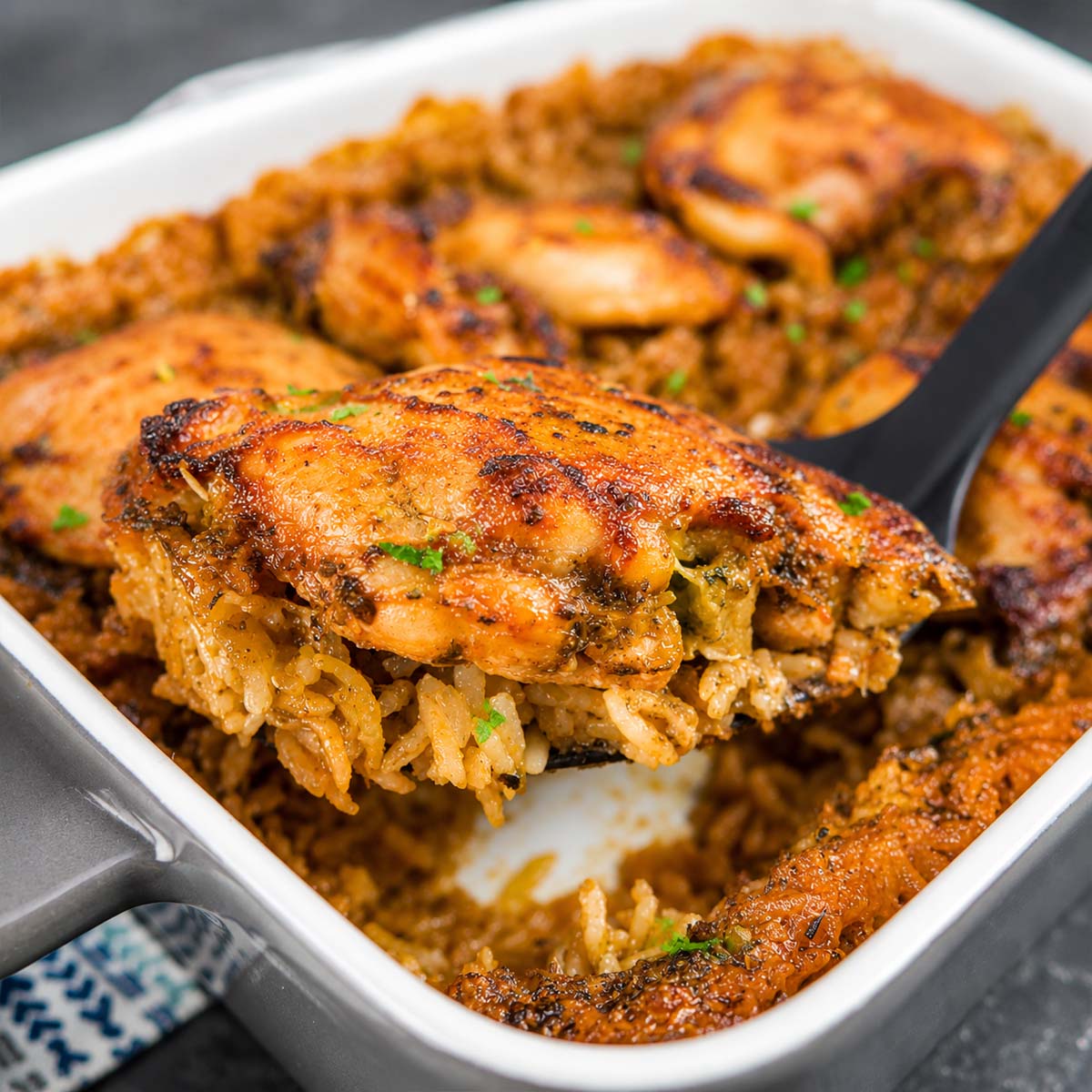 Baked chicken thighs and rice casserole in a white dish with golden, seasoned chicken pieces on top and parsley garnish. close up of the chicken thigh and rice scooped with a large spoon.
