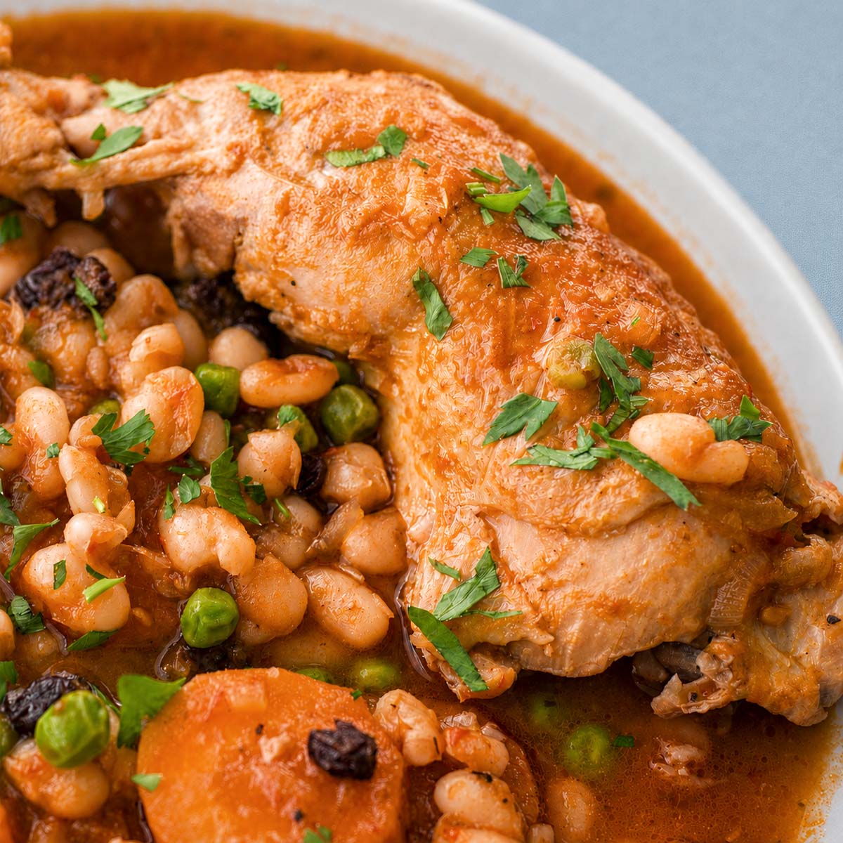 Close-up of chicken and bean stew with carrots, peas, and raisins in a tomato sauce, garnished with fresh parsley.