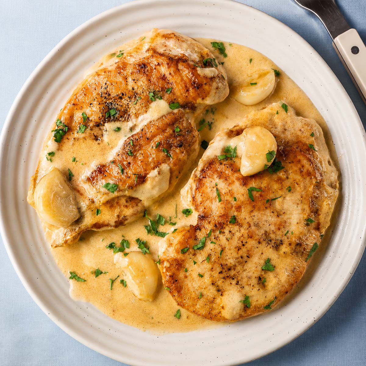 Overhead view of one-pan creamy garlic chicken simmering in a rich garlic cream sauce with golden seared chicken breasts and whole garlic cloves.