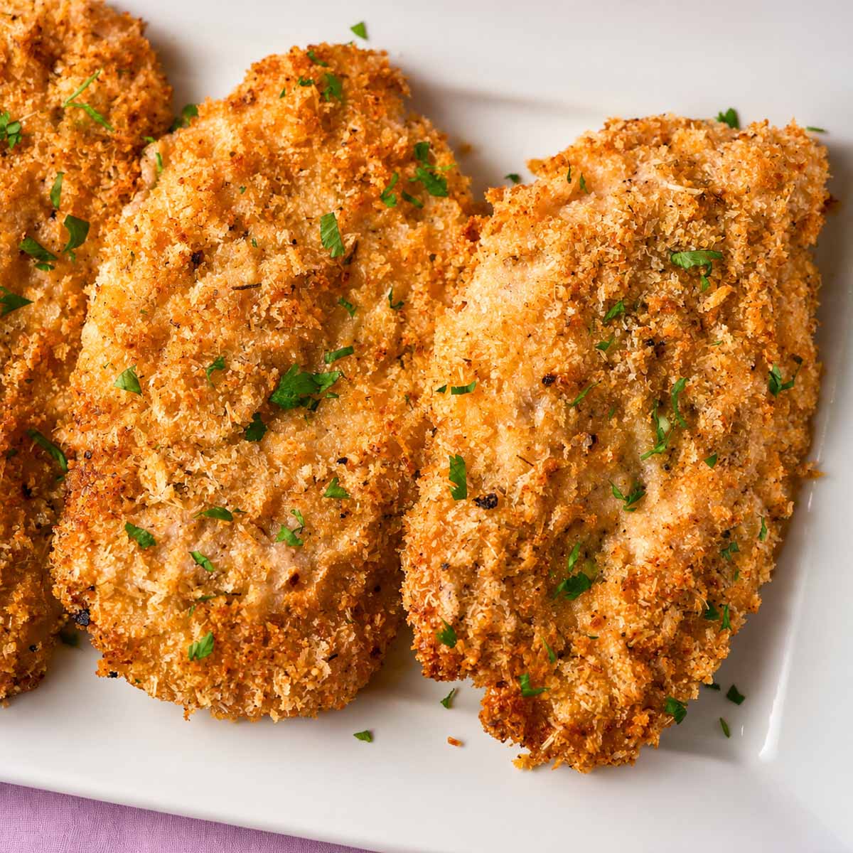 Crispy baked Parmesan crusted chicken on a white platter with a golden panko and Parmesan coating, garnished with fresh parsley.