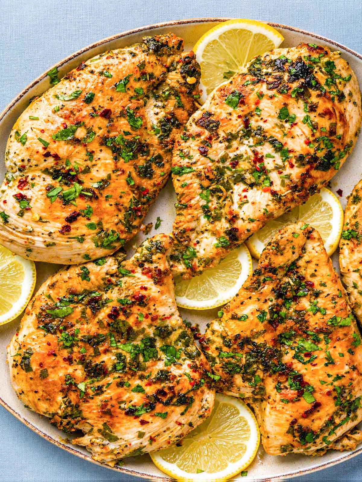 Close-up of Greek lemon chicken cutlets in a skillet with chopped parsley, red pepper flakes, and lemon slices.