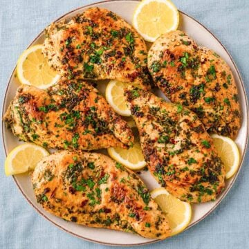 Greek lemon chicken cutlets in a skillet topped with chopped parsley and lemon slices.
