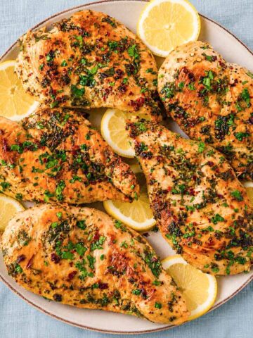 Greek lemon chicken cutlets in a skillet topped with chopped parsley and lemon slices.