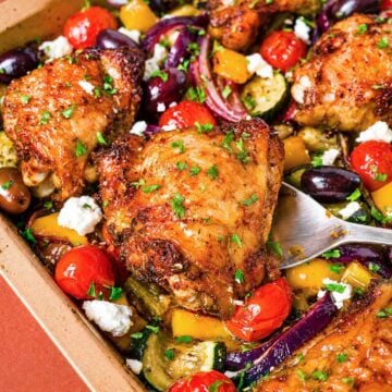 Greek sheet pan chicken thighs in a baking dish with roasted zucchini, red onion, cherry tomatoes, Kalamata olives, feta, and herbs.