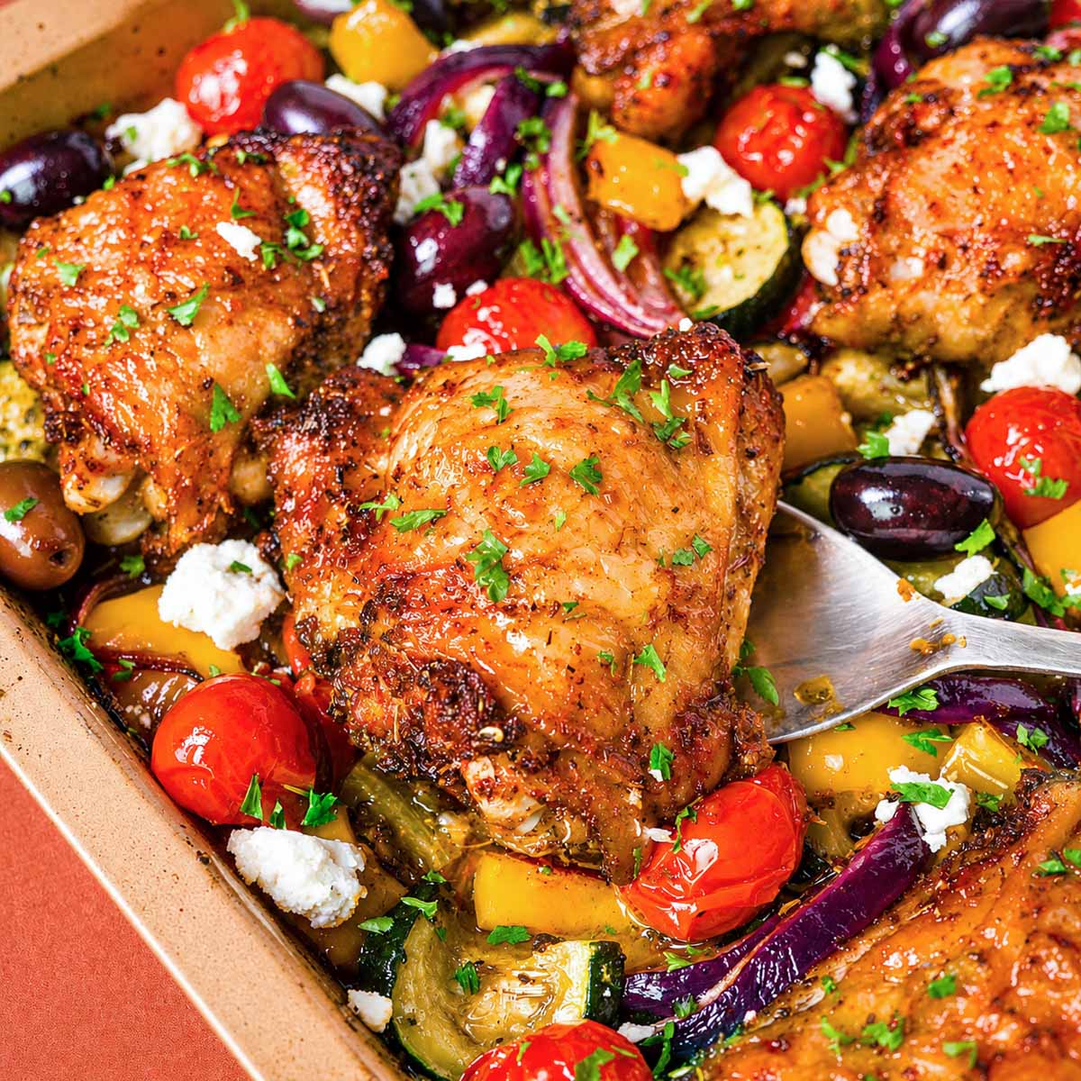 Greek sheet pan chicken thighs in a baking dish with roasted zucchini, red onion, cherry tomatoes, Kalamata olives, feta, and herbs.