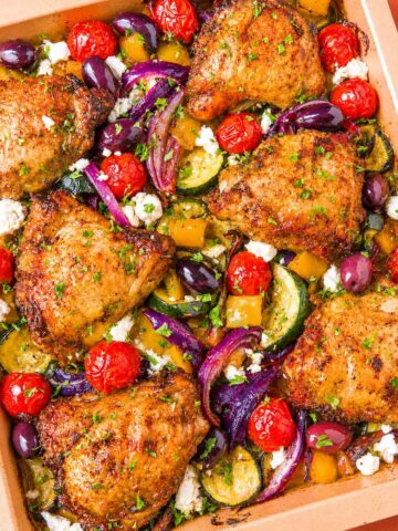 Greek sheet pan chicken thighs with roasted zucchini, red onion, cherry tomatoes, Kalamata olives, and crumbled feta in a baking dish.