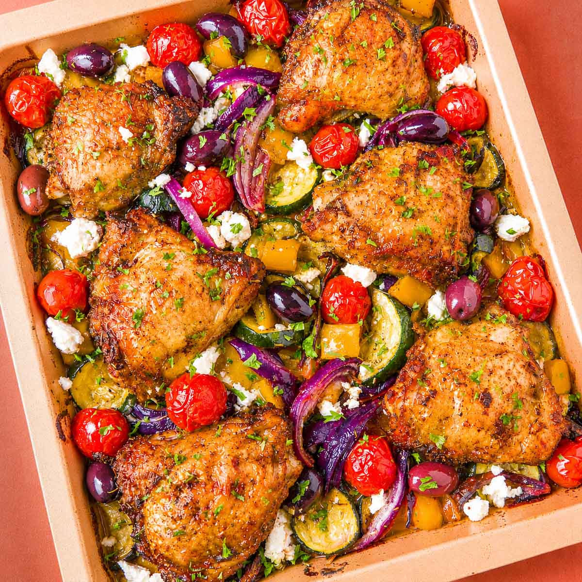 Greek sheet pan chicken thighs with roasted zucchini, red onion, cherry tomatoes, Kalamata olives, and crumbled feta in a baking dish.