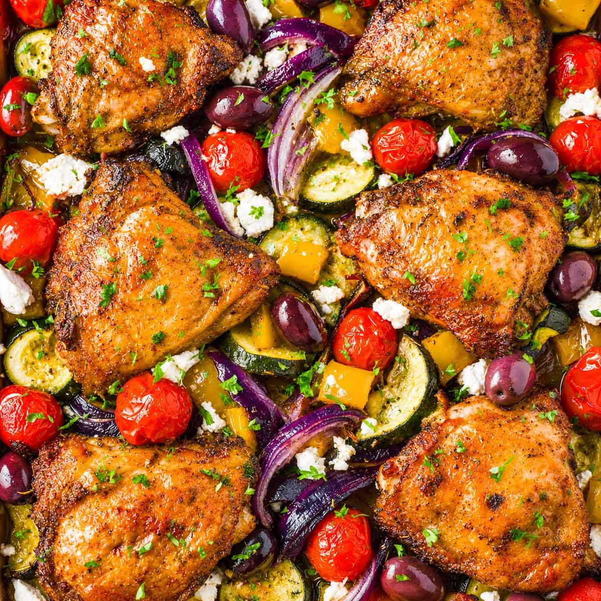 Close-up of Greek sheet pan chicken thighs with roasted zucchini, red onion, cherry tomatoes, Kalamata olives, and feta.