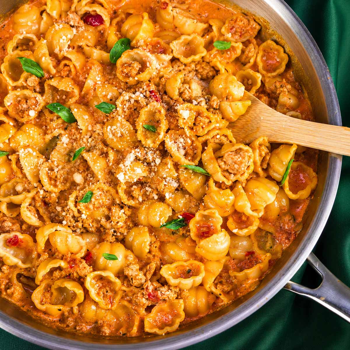 Ground turkey pasta shells in a creamy tomato sauce in a stainless steel skillet, topped with Parmesan and basil, with a wooden spoon stirring the pasta.