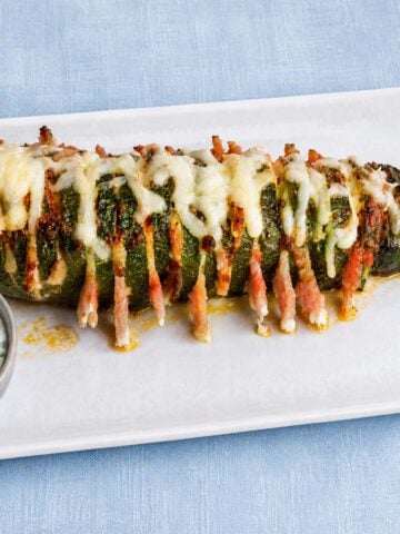 Hasselback zucchini baked with melted cheese and bacon, served on a white plate with creamy herb sauce