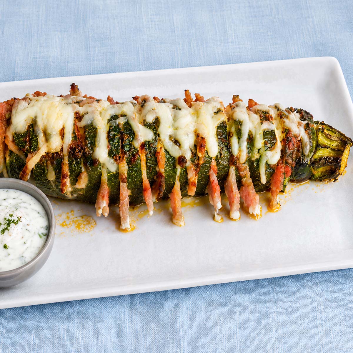 Hasselback zucchini baked with melted cheese and bacon, served on a white plate with creamy herb sauce