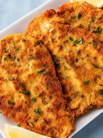 Crispy Italian breaded chicken cutlets on a white platter, garnished with fresh parsley and served with lemon wedges.