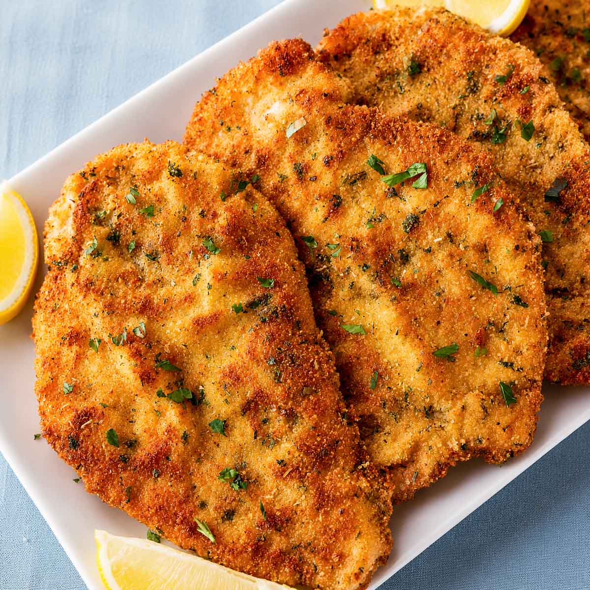 Crispy Italian breaded chicken cutlets on a white platter, garnished with fresh parsley and served with lemon wedges. 