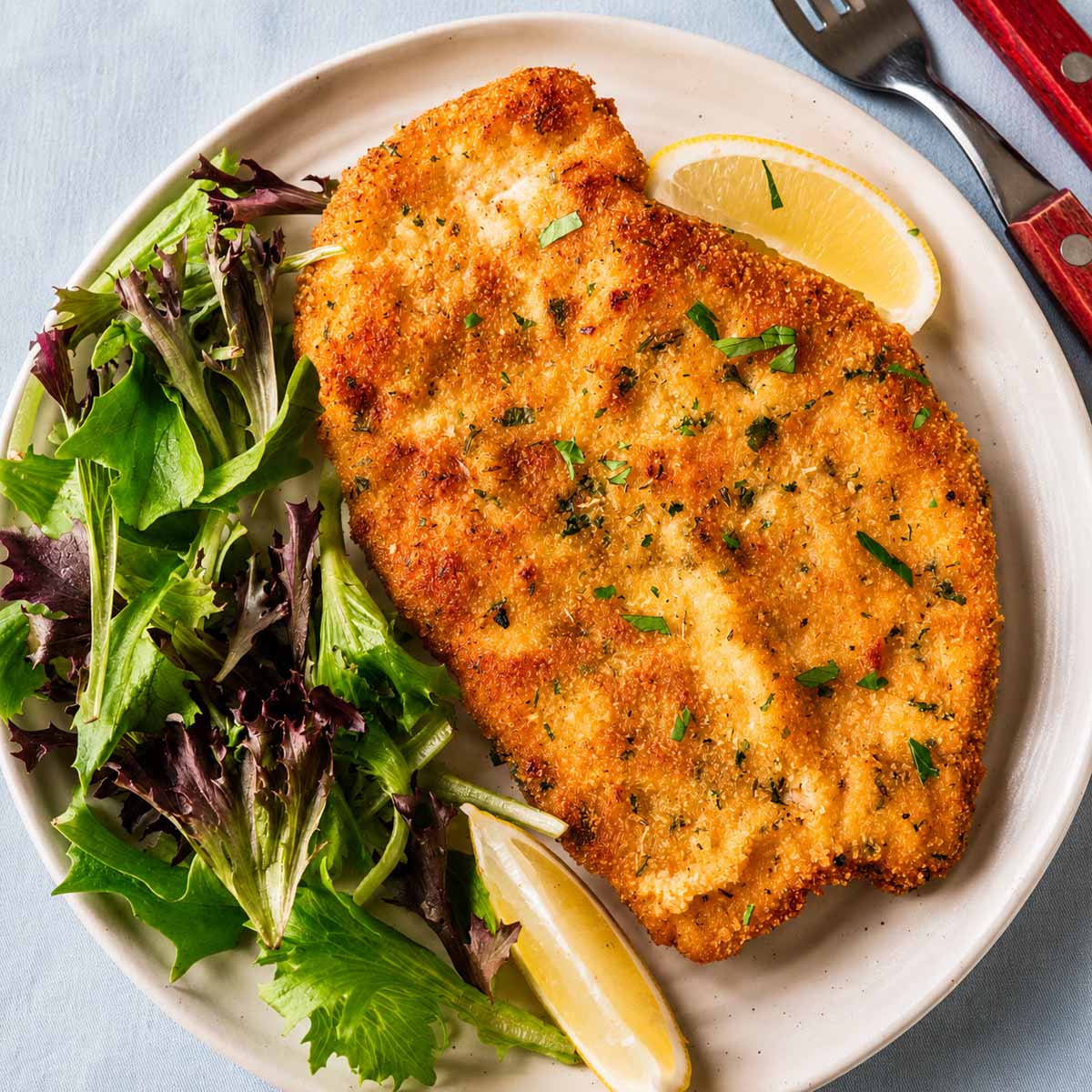 Crispy Italian breaded chicken cutlet on a plate with mixed greens and lemon wedges. 