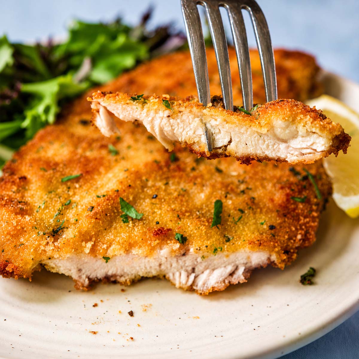 Fork holding a bite of crispy Italian breaded chicken cutlet, showing the juicy inside, with more cutlet, greens, and a lemon wedge on the plate below. 