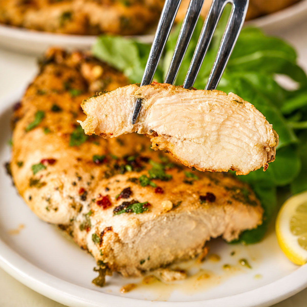 Fork holding a slice of juicy lemon garlic chicken breast over a plate with the remaining chicken and a green salad in the background.