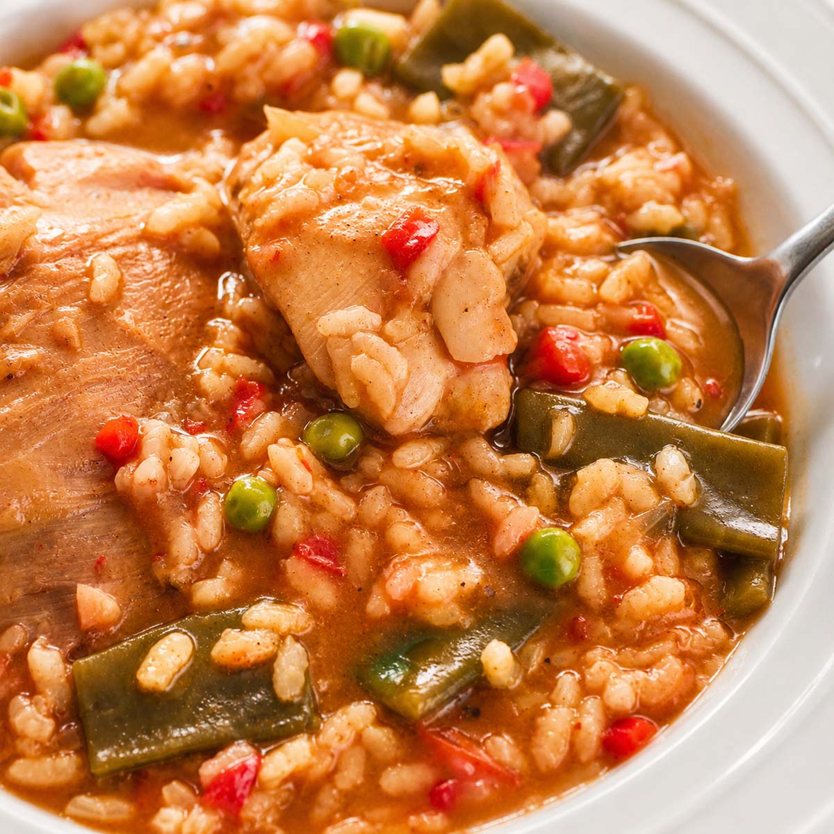 Spoon scooping one pot Spanish chicken and rice with tender chicken, peas, and green beans in a tomato-based broth. 