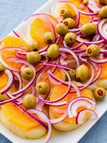 Spanish potato salad with orange slices, green olives, and thinly sliced red onion on a white serving platter.