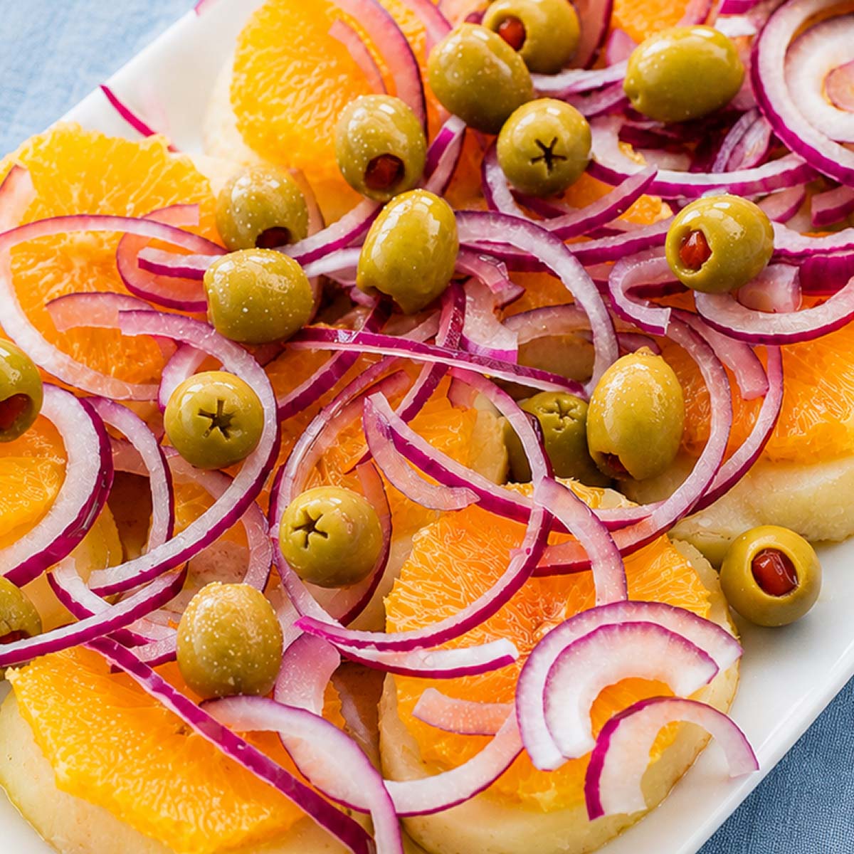 Close up of Spanish potato salad with orange slices, green olives, and thinly sliced red onion on a white serving platter.
