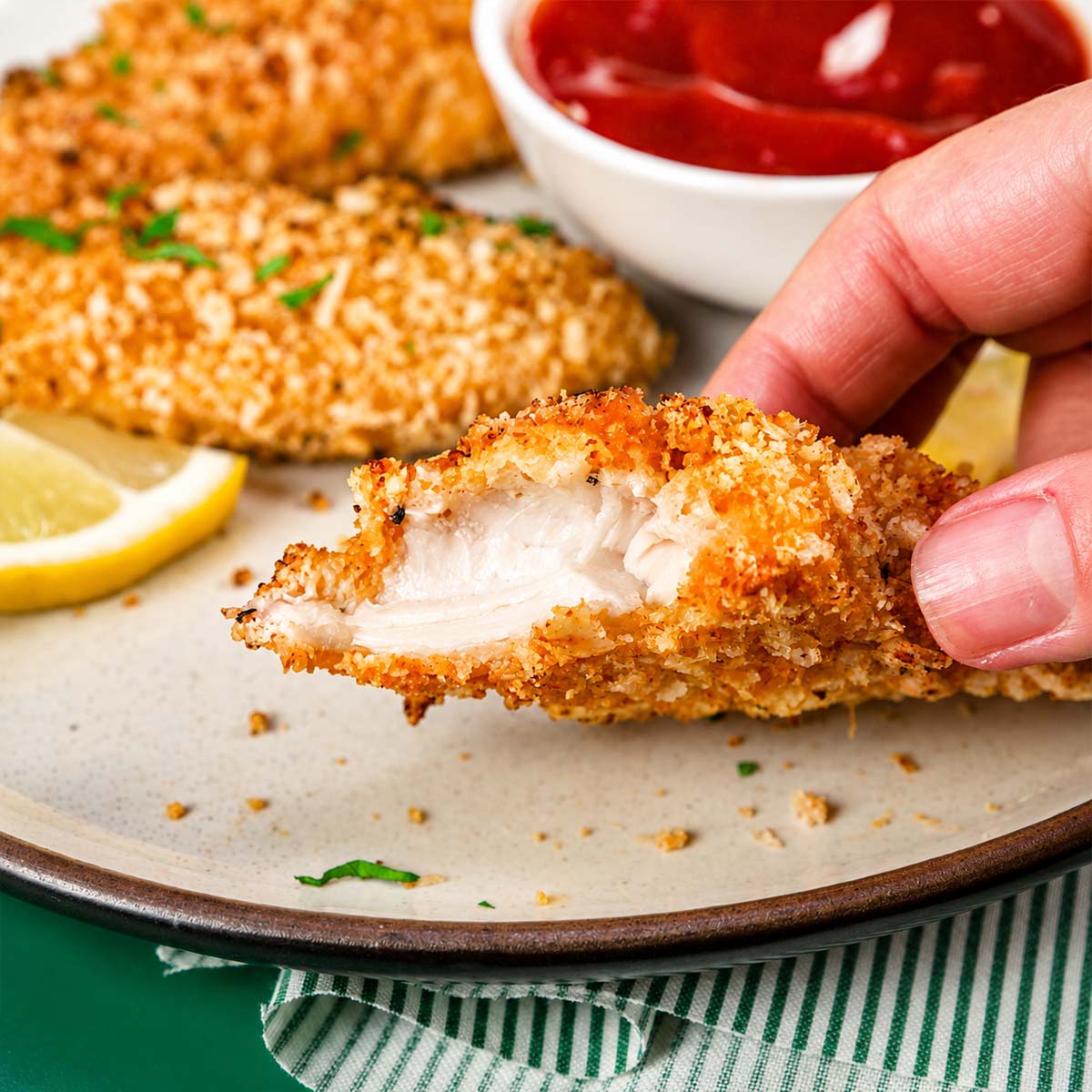 Hand holding a bitten oven-baked chicken tender showing the juicy inside, with crispy chicken tenders, ketchup dipping sauce, and a lemon wedge in the background.