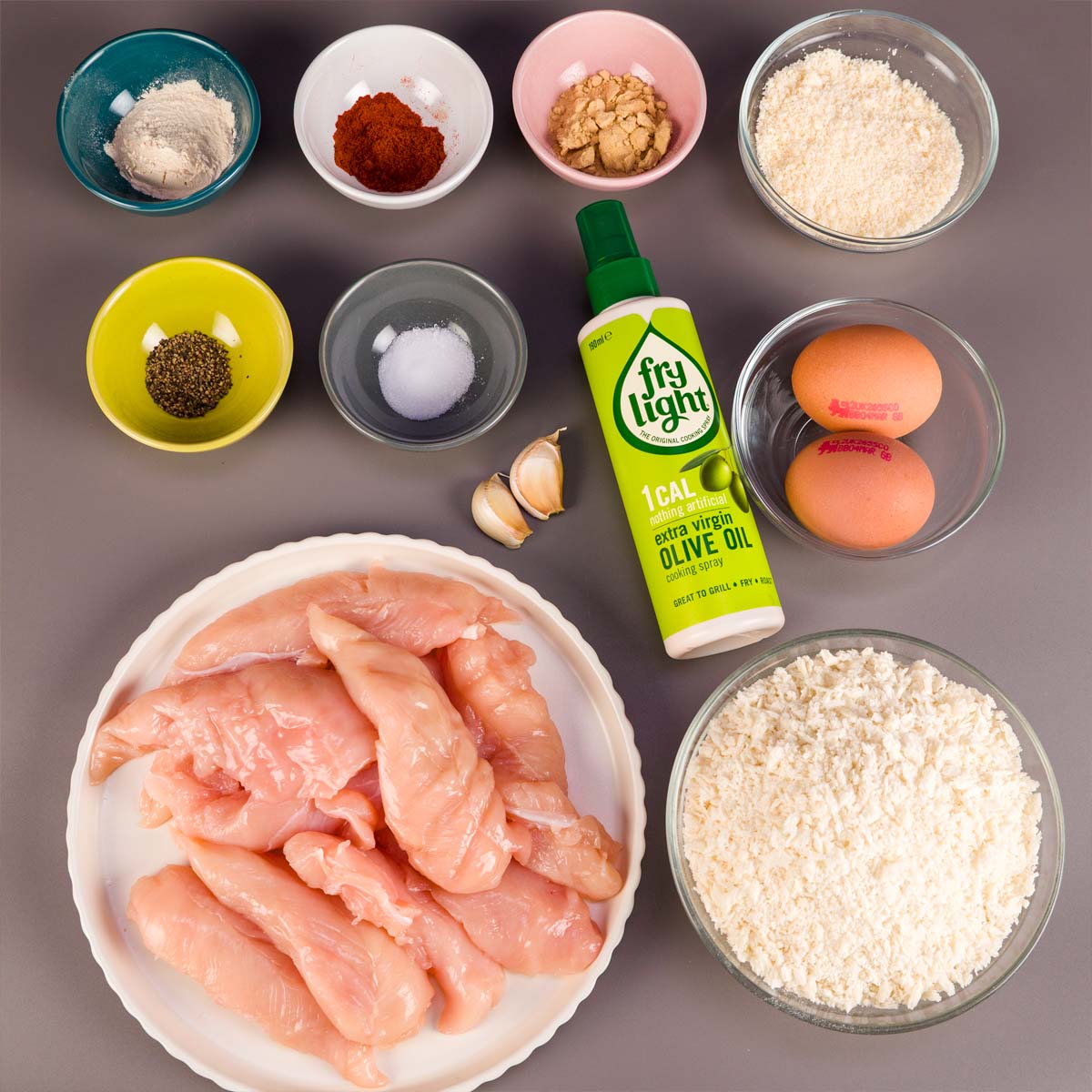 Ingredients for oven-baked chicken tenders laid out on a gray surface, including raw chicken tenders, flour, panko breadcrumbs, grated Parmesan, eggs, garlic cloves, salt, pepper, paprika, garlic powder, and olive oil cooking spray.