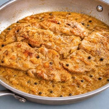 Pork chops in peppercorn sauce cooking in a skillet with onions and whole black peppercorns